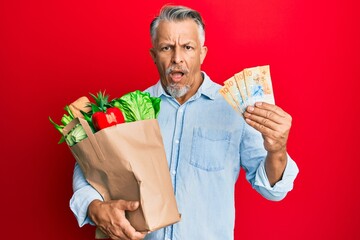 Middle age grey-haired man holding groceries and swiss franc banknotes in shock face, looking...
