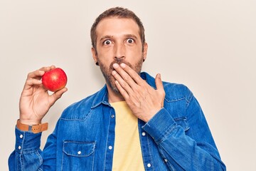 Young handsome man holding red apple covering mouth with hand, shocked and afraid for mistake....