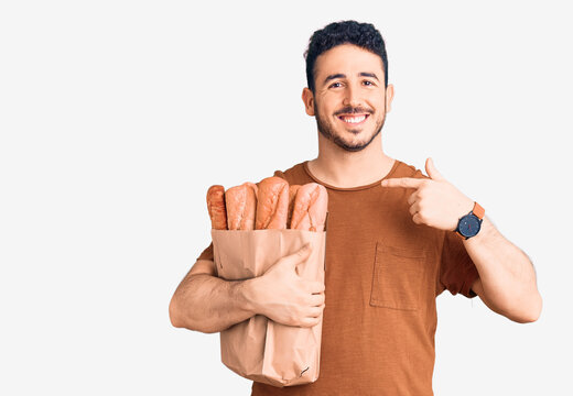 Young hispanic man holding paper bag with bread pointing finger to one self smiling happy and proud