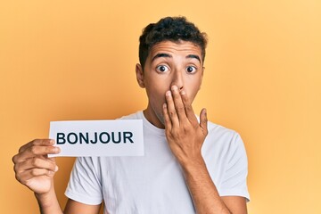 Young handsome african american man holding bonjour french greeting word covering mouth with hand, shocked and afraid for mistake. surprised expression © Krakenimages.com