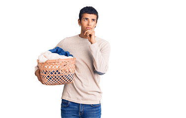 Young african amercian man holding laundry basket serious face thinking about question with hand on...