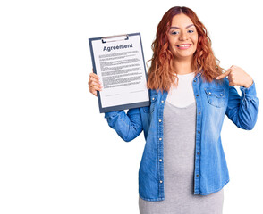 Young latin woman holding clipboard with agreement document pointing finger to one self smiling happy and proud