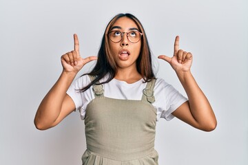 Young latin woman wearing casual clothes and glasses amazed and surprised looking up and pointing with fingers and raised arms.