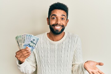 Handsome hispanic man with beard holding dollars celebrating achievement with happy smile and winner expression with raised hand