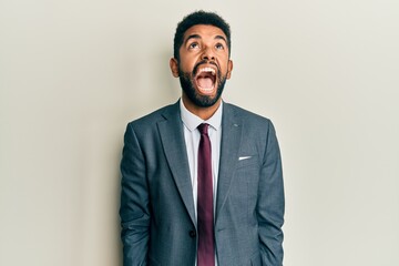 Handsome hispanic man with beard wearing business suit and tie angry and mad screaming frustrated and furious, shouting with anger. rage and aggressive concept.