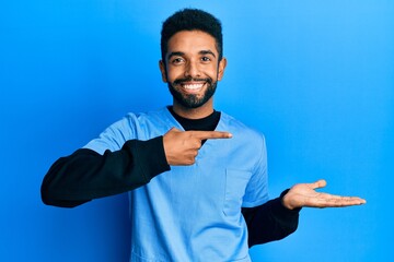 Handsome hispanic man with beard wearing blue male nurse uniform amazed and smiling to the camera while presenting with hand and pointing with finger.