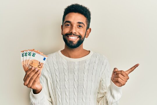 Handsome hispanic man with beard holding bunch of 50 euro banknotes smiling happy pointing with hand and finger to the side