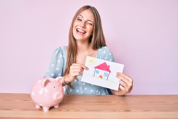 Young blonde girl holding house draw and piggy bank winking looking at the camera with sexy...