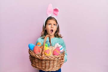 Obraz na płótnie Canvas Little beautiful girl wearing cute easter bunny ears holding wicker basket with colored eggs angry and mad screaming frustrated and furious, shouting with anger looking up.