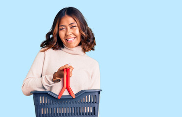 Young beautiful mixed race woman holding supermarket shopping basket looking positive and happy standing and smiling with a confident smile showing teeth