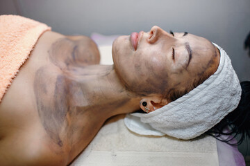 Young beautiful woman in spa salon with black mask on her face, master beautician applied a mask on the face of a young girl to cleanse the pores of the skin. Facial skin care in modern cosmetology