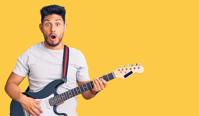 Handsome latin american young man playing electric guitar scared and amazed with open mouth for surprise, disbelief face