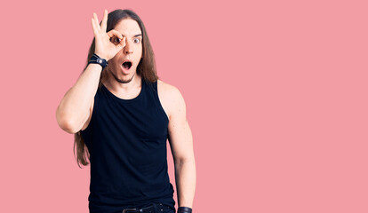 Young adult man with long hair wearing goth style with black clothes doing ok gesture shocked with surprised face, eye looking through fingers. unbelieving expression.