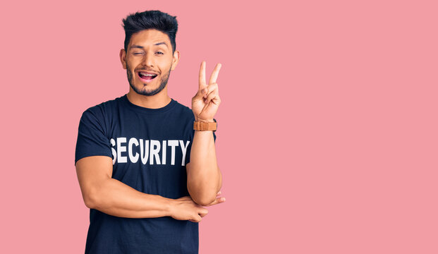 Handsome latin american young man wearing security t shirt smiling with happy face winking at the camera doing victory sign with fingers. number two.