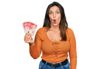 Beautiful hispanic woman holding 20 israel shekels banknotes scared and amazed with open mouth for...