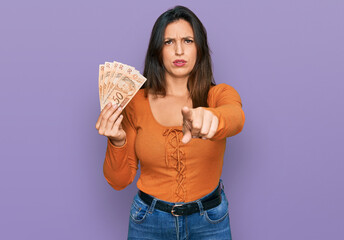 Beautiful hispanic woman holding 50 brazilian real banknotes pointing with finger to the camera and to you, confident gesture looking serious