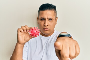 Young latin man holding virus toy pointing with finger to the camera and to you, confident gesture looking serious