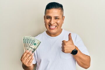 Young latin man holding 20 dollars banknotes smiling happy and positive, thumb up doing excellent and approval sign