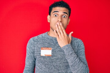 Young handsome hispanic man wearing hello my name is sticker identification covering mouth with hand, shocked and afraid for mistake. surprised expression