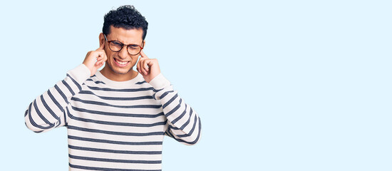 Hispanic handsome young man wearing casual clothes and glasses covering ears with fingers with annoyed expression for the noise of loud music. deaf concept.