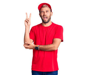 Young handsome blond man wearing t-shirt and cap smiling with happy face winking at the camera doing victory sign. number two.