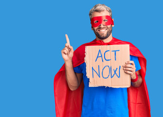 Young blond man wearing super hero costume holding act now cardboard banner surprised with an idea or question pointing finger with happy face, number one