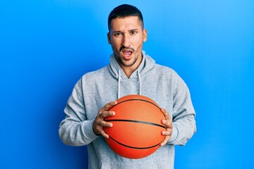 Handsome man with tattoos holding basketball ball in shock face, looking skeptical and sarcastic, surprised with open mouth