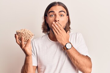 Young handsome man holding chickpeas bowl covering mouth with hand, shocked and afraid for mistake....