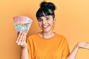 Young hispanic girl holding 100 new zealand dollars banknote celebrating achievement with happy smile and winner expression with raised hand