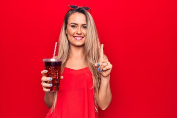 Beautiful blonde woman drinking cola beverage refreshment using straw over red background smiling with an idea or question pointing finger with happy face, number one