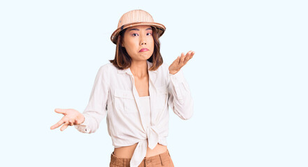 Young beautiful chinese girl wearing explorer hat clueless and confused expression with arms and...