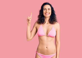 Naklejka premium Young beautiful hispanic woman wearing bikini showing and pointing up with finger number one while smiling confident and happy.