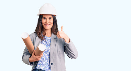 Young beautiful brunette woman wearing architect hardhat holding blueprint smiling happy and positive, thumb up doing excellent and approval sign
