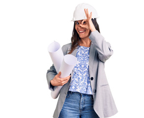 Young beautiful brunette woman wearing architect hardhat holding blueprint smiling happy doing ok sign with hand on eye looking through fingers