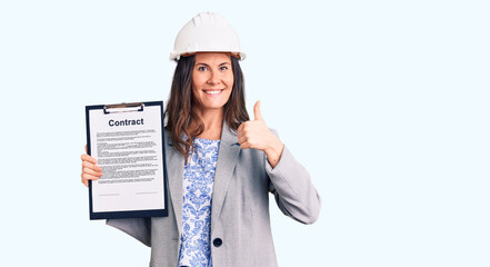 Young beautiful brunette woman wearing architect hardhat holding contract smiling happy and positive, thumb up doing excellent and approval sign
