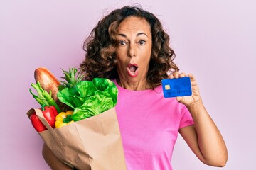 Middle age hispanic woman holding groceries and credit card afraid and shocked with surprise and amazed expression, fear and excited face.