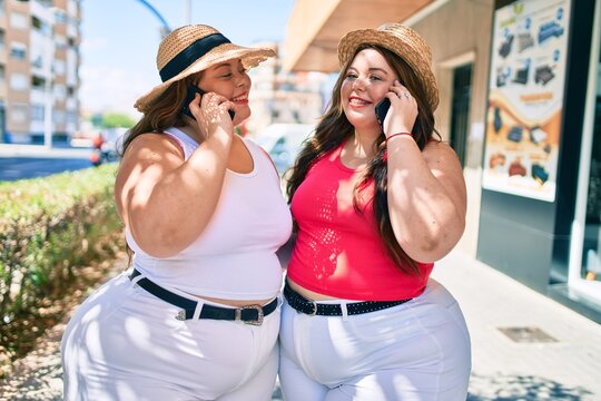 Two plus size overweight sisters twins women speaking on the phone outdoors on a sunny day