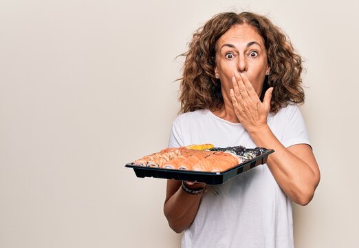 Middle age beautiful woman holding takeaway tray with sushi over isolated white background covering mouth with hand, shocked and afraid for mistake. Surprised expression