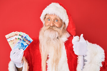 Old senior man with grey hair and long beard wearing santa claus costume holding canadian dollars smiling happy and positive, thumb up doing excellent and approval sign