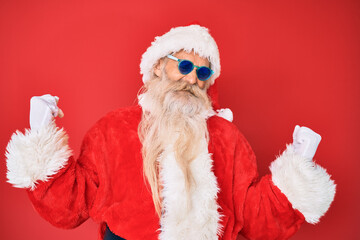 Old senior man wearing santa claus costume and sunglasses very happy and excited doing winner gesture with arms raised, smiling and screaming for success. celebration concept.