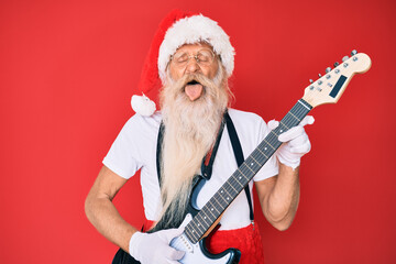 Old senior man wearing santa claus costume playing electric guitar sticking tongue out happy with funny expression. © Krakenimages.com