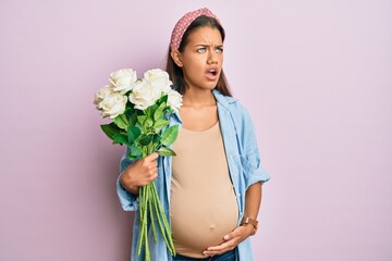 Beautiful hispanic woman expecting a baby holding flowers angry and mad screaming frustrated and furious, shouting with anger. rage and aggressive concept.