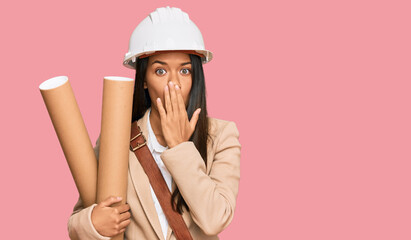 Beautiful hispanic woman holding paper blueprints covering mouth with hand, shocked and afraid for...