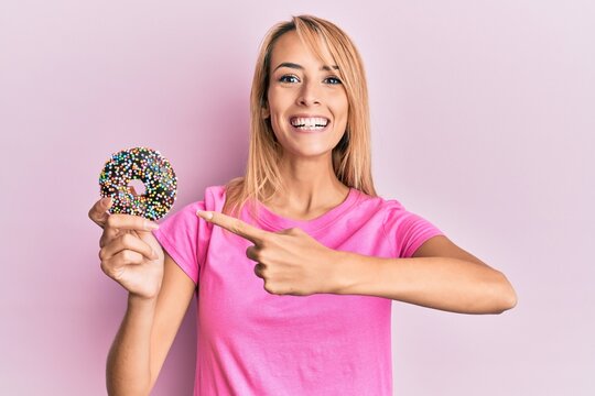 Beautiful blonde woman holding donut smiling happy pointing with hand and finger