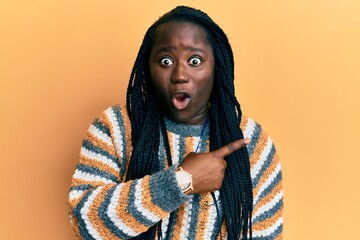 Young black woman with braids wearing casual winter sweater surprised pointing with finger to the side, open mouth amazed expression.