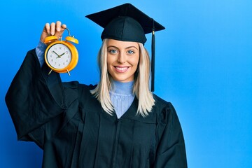 Beautiful blonde woman wearing graduation cap and ceremony robe holding alarm clock looking...