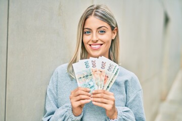 Young blonde girl smiling happy holding czech koruna banknotes at the city.