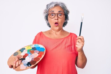 Senior hispanic grey- haired woman holding paintbrush and palette scared and amazed with open mouth...