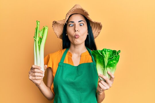Beautiful young woman wearing gardener apron holding vegetables making fish face with mouth and squinting eyes, crazy and comical.