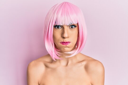 Young man wearing woman make up wearing pink wig puffing cheeks with funny face. mouth inflated with air, crazy expression.